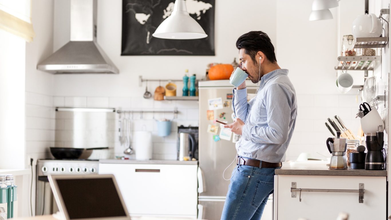 A man in a denim shirt and jeans standing in a bright kitchen, drinking from a mug and listening to a digital marketing podcast