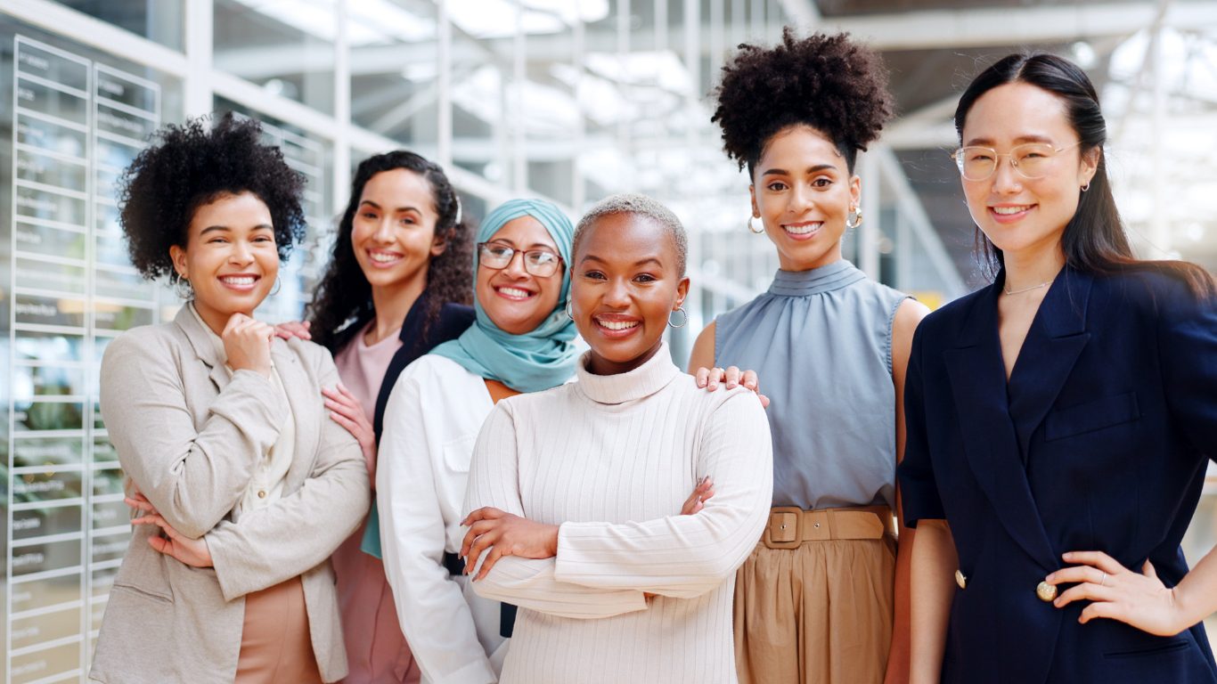 Group of six diverse women entrepreneurs, who are fractional cmos and entrepreneurs, smiling at the camera, standing in a well-lit modern office environment. 